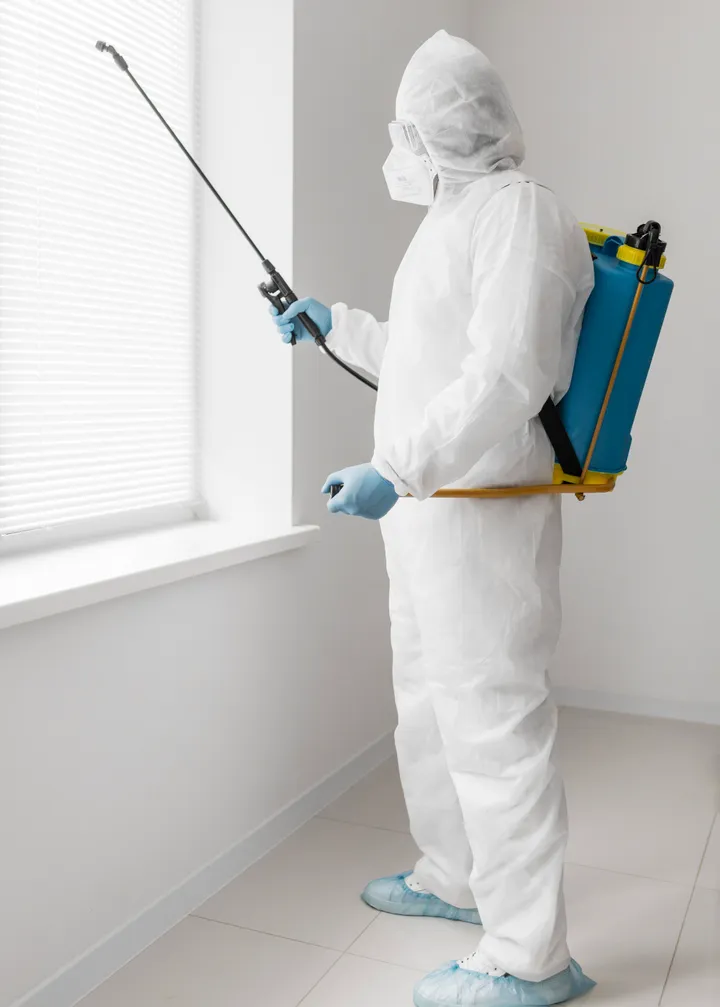Técnico con traje de protección, gafas y tapabocas aplicando tratamiento contra cucarachas con fumigadora de espalda